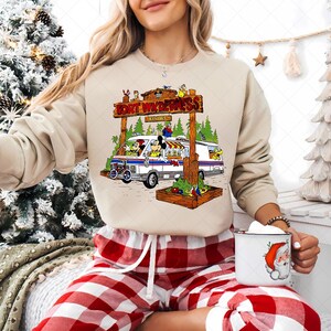 K&ouml;nnte beinhalten: Beiges Sweatshirt mit einer bunten Grafik eines Vans mit Zeichentrickfiguren und dem Text "FORT WILDERNESS RESORT". Der Van parkt vor einem Holzschild. Die Person tr&auml;gt eine rot-wei&szlig; karierte Pyjamahose und h&auml;lt eine Tasse mit einem Weihnachtsmann-Design.