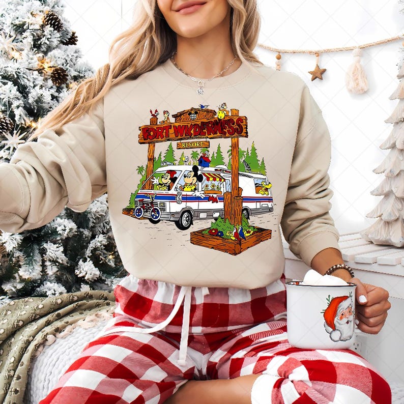 K&ouml;nnte beinhalten: Beiges Sweatshirt mit einer bunten Grafik eines Vans mit Zeichentrickfiguren und dem Text "FORT WILDERNESS RESORT". Der Van parkt vor einem Holzschild. Die Person tr&auml;gt eine rot-wei&szlig; karierte Pyjamahose und h&auml;lt eine Tasse mit einem Weihnachtsmann-Design.