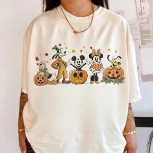 May include: An off-white t-shirt featuring a graphic print of Disney characters including Donald Duck, Goofy, Mickey Mouse, Minnie Mouse, and Pluto. The characters are depicted with pumpkins and autumn leaves in a festive, seasonal design.
