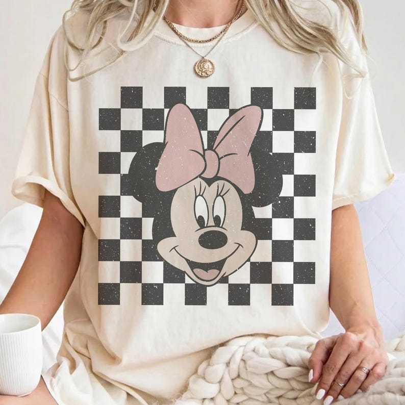 May include: Cream-colored t-shirt featuring a Minnie Mouse graphic. The design includes a black and white checkered background and a pink bow. The graphic tee is a casual wear item.