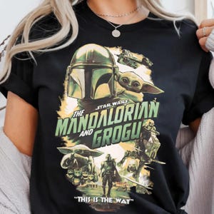 May include: Black t-shirt featuring a Star Wars design with The Mandalorian and Grogu. The graphic includes images of the Mandalorian, Grogu, and various vehicles and characters from the series. Text on the shirt reads "The Mandalorian and Grogu" and "This is the Way."