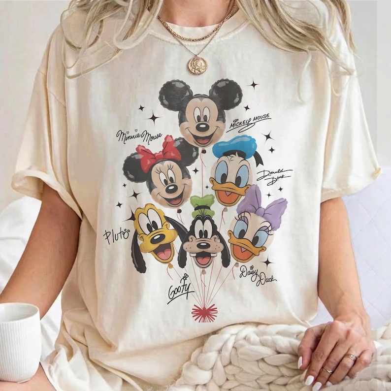 May include: Cream-colored t-shirt featuring a graphic of Mickey Mouse, Minnie Mouse, Donald Duck, Daisy Duck, Pluto, and Goofy as balloon characters. The shirt includes the characters' names and the words "Mickey Mouse".