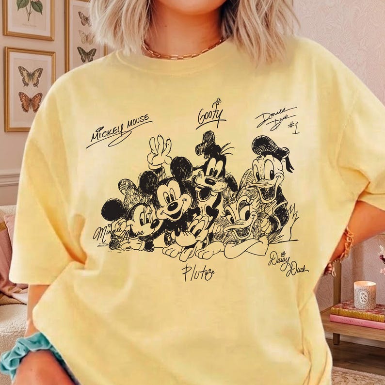 Pu&ograve; includere: T-shirt giallo chiaro con un'illustrazione in bianco e nero di Topolino, Minnie, Pippo, Paperino, Paperina e Pluto. I nomi dei personaggi sono scritti sopra o sotto. La maglietta &egrave; di colore giallo chiaro.