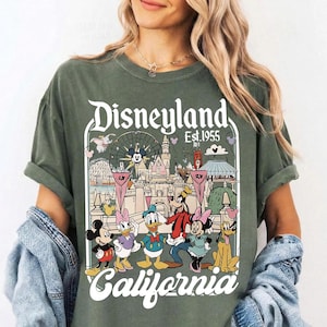 May include: Olive green t-shirt featuring a Disneyland graphic with Mickey Mouse, Minnie Mouse, Donald Duck, Goofy, and Daisy Duck. The graphic includes the text "Disneyland Est. 1955 California". The shirt has rolled-up sleeves.