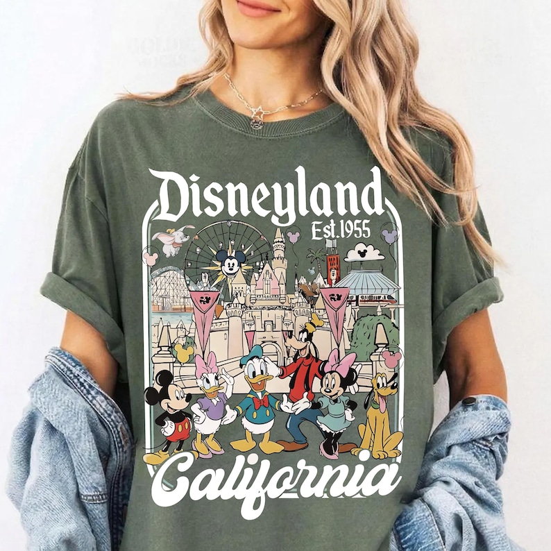 May include: Olive green t-shirt featuring a Disneyland graphic with Mickey Mouse, Minnie Mouse, Donald Duck, Goofy, and Daisy Duck. The graphic includes the text "Disneyland Est. 1955 California". The shirt has rolled-up sleeves.
