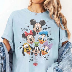 May include: Light blue t-shirt featuring a graphic of Disney characters as balloons, including Mickey Mouse, Minnie Mouse, Donald Duck, Daisy Duck, Goofy, and Pluto. The names of the characters are written in cursive. The shirt is a light blue color.