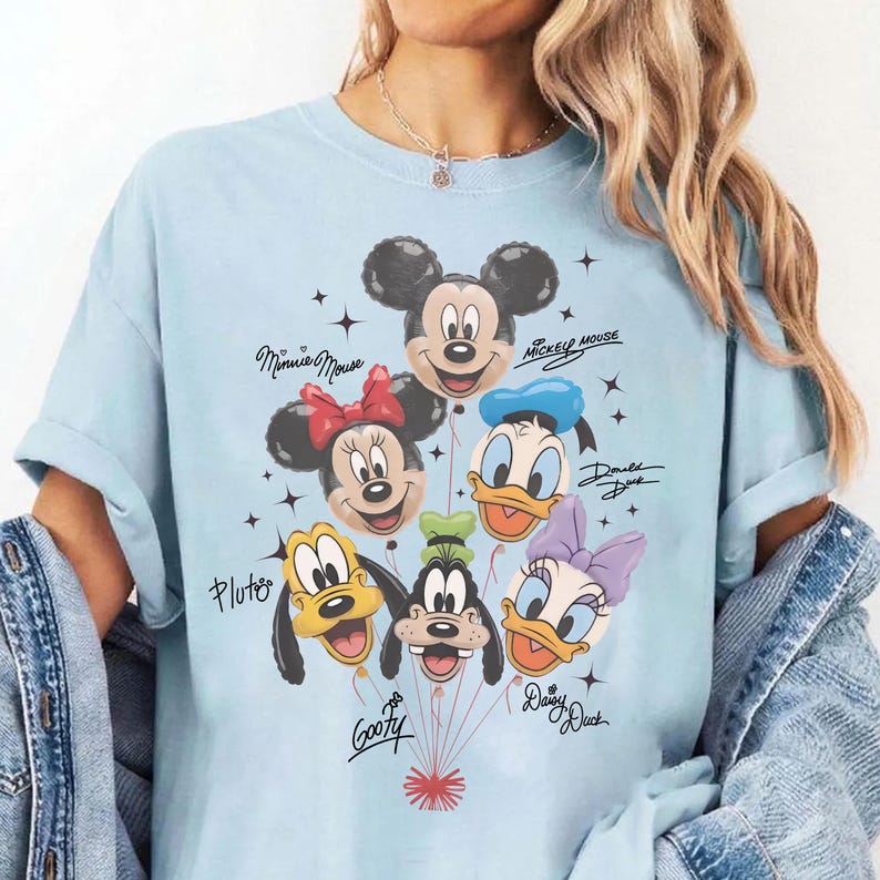May include: Light blue t-shirt featuring a graphic of Disney characters as balloons, including Mickey Mouse, Minnie Mouse, Donald Duck, Daisy Duck, Goofy, and Pluto. The names of the characters are written in cursive. The shirt is a light blue color.