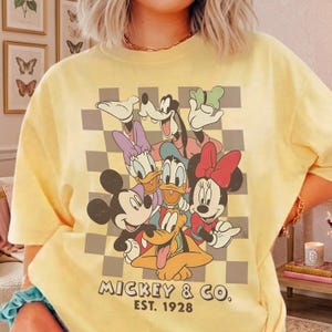 May include: Yellow t-shirt featuring a graphic of Mickey Mouse, Minnie Mouse, Donald Duck, Daisy Duck, Goofy, and Pluto. The design includes a checkered background and the text "Mickey & Co. Est. 1928".