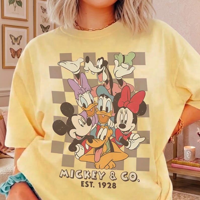 May include: Yellow t-shirt featuring a graphic of Mickey Mouse, Minnie Mouse, Donald Duck, Daisy Duck, Goofy, and Pluto. The design includes a checkered background and the text "Mickey & Co. Est. 1928".