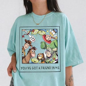 May include: Light blue t-shirt featuring a framed graphic of Toy Story characters, including Woody, Buzz Lightyear, and Rex. The text "YOU'VE GOT A FRIEND IN ME" is printed below the graphic. The shirt is made of a soft material.