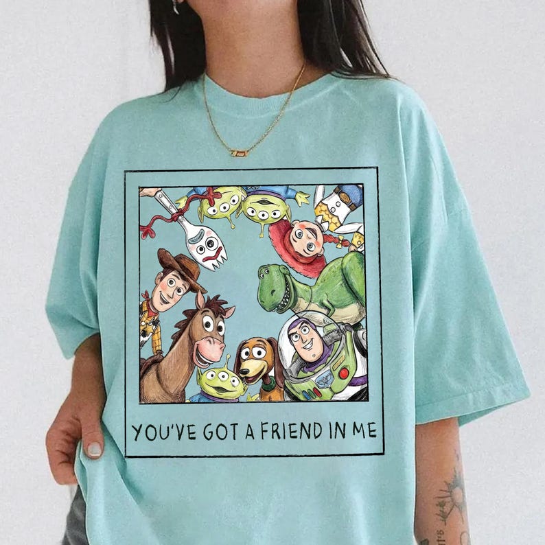 May include: Light blue t-shirt featuring a framed graphic of Toy Story characters, including Woody, Buzz Lightyear, and Rex. The text "YOU'VE GOT A FRIEND IN ME" is printed below the graphic. The shirt is made of a soft material.