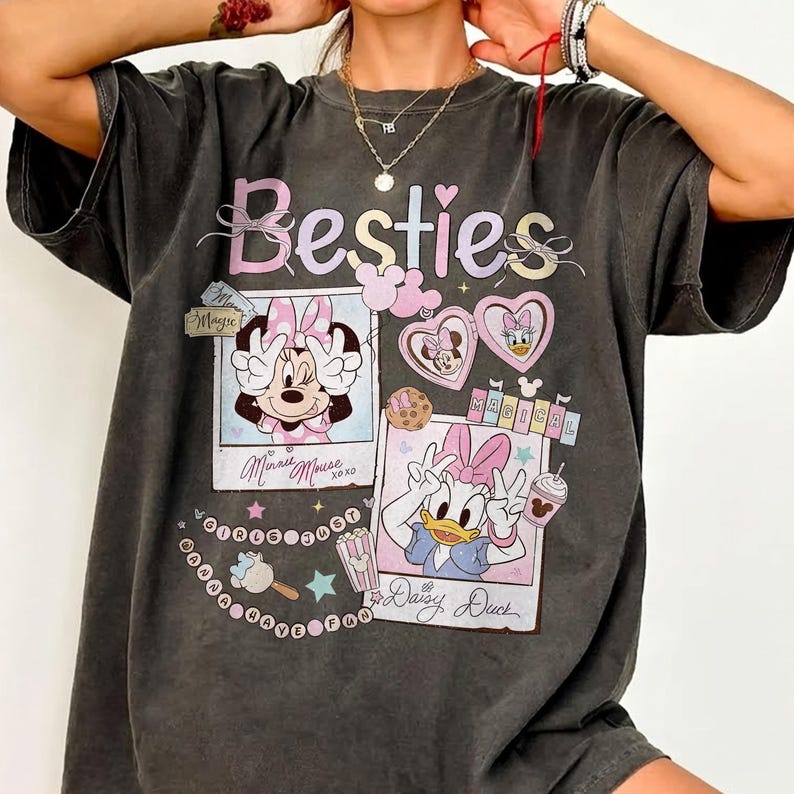 Peut inclure: T-shirt gris fonc&eacute; avec le mot "Besties" en rose et violet, et des graphiques de Minnie Mouse et Daisy Duck. Le t-shirt comporte les textes "Minnie Mouse" et "Daisy Duck" sous les graphiques, ainsi que d'autres &eacute;l&eacute;ments d&eacute;coratifs.