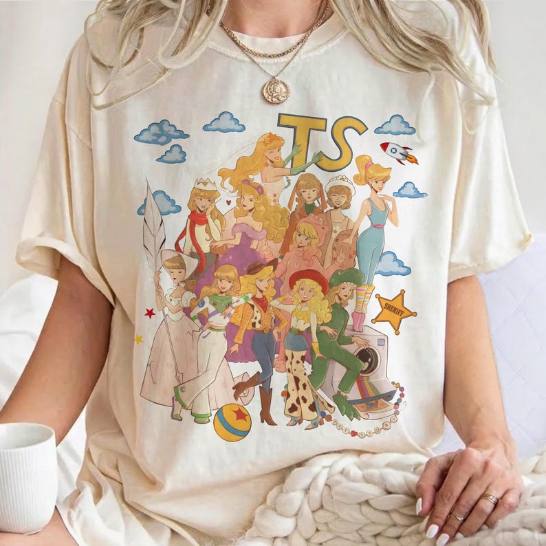 May include: A cream-colored short-sleeve t-shirt featuring a large graphic print of multiple animated female characters in various outfits. The design includes the letters TS, a rocket, a yellow star badge labeled SHERIFF, and a colorful ball.