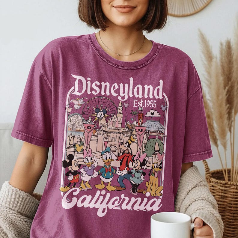 May include: A maroon t-shirt featuring a Disneyland California graphic. The design includes Mickey Mouse, Minnie Mouse, Donald Duck, Daisy Duck, Goofy, and Pluto, along with the Disneyland logo and the text "Est.1955".