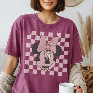 May include: A maroon t-shirt featuring a Minnie Mouse graphic. The design includes a checkered background in pink and white, with Minnie's face and bow prominently displayed. The shirt has short sleeves and a crew neck.