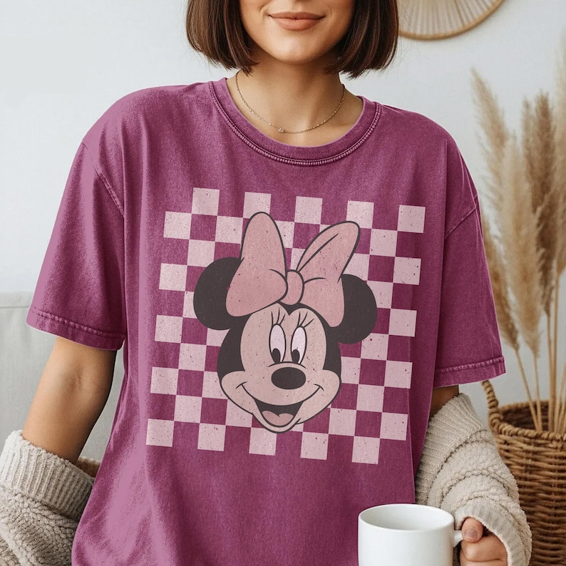 May include: A maroon t-shirt featuring a Minnie Mouse graphic. The design includes a checkered background in pink and white, with Minnie's face and bow prominently displayed. The shirt has short sleeves and a crew neck.