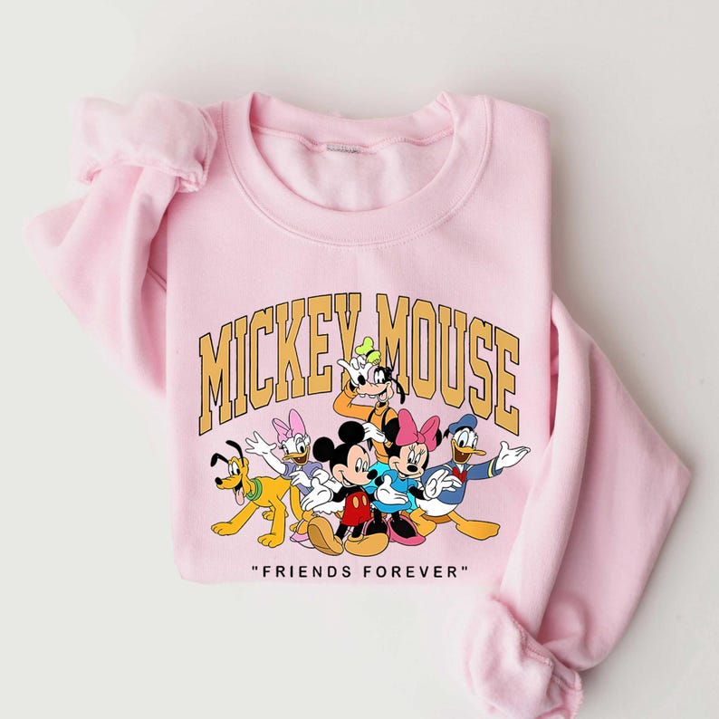 May include: A pale pink sweatshirt with a cartoon graphic of Mickey Mouse and friends. The words "MICKEY MOUSE" are arched across the top in gold lettering, with "FRIENDS FOREVER" below. The cartoon characters include Mickey, Minnie, Donald, and Goofy.