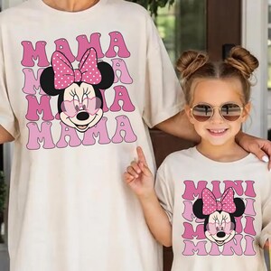 K&ouml;nnte beinhalten: Zwei passende cremefarbene T-Shirts. Das Erwachsenen-Shirt zeigt eine Minnie-Maus-Grafik mit dem Wort "MAMA" in rosa Buchstaben. Das Kinder-Shirt hat ein &auml;hnliches Design mit dem Wort "MINI". Beide Shirts haben eine Cartoon-Minnie-Maus mit einer rosa Schleife.