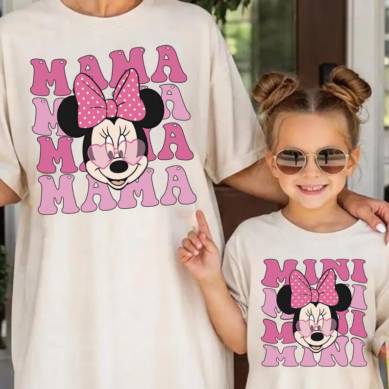 K&ouml;nnte beinhalten: Zwei passende cremefarbene T-Shirts. Das Erwachsenen-Shirt zeigt eine Minnie-Maus-Grafik mit dem Wort "MAMA" in rosa Buchstaben. Das Kinder-Shirt hat ein &auml;hnliches Design mit dem Wort "MINI". Beide Shirts haben eine Cartoon-Minnie-Maus mit einer rosa Schleife.