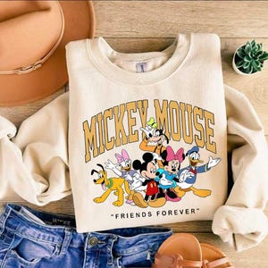 May include: A cream-coloured sweatshirt with a graphic of Mickey Mouse and friends, and the text "MICKEY MOUSE" in gold. Below the graphic, the text reads "FRIENDS FOREVER". The sweatshirt is shown with blue jeans and a brown hat.