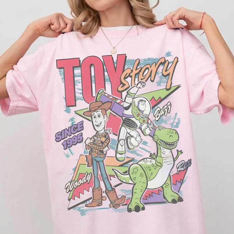 May include: A light pink t-shirt featuring a graphic of characters from the movie Toy Story. The design includes Woody, Buzz Lightyear, and Rex, with the words "TOY STORY" and "SINCE 1995".