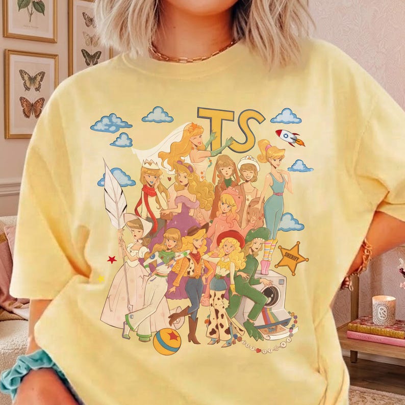May include: A pale yellow short-sleeve t-shirt featuring a graphic print of various cartoon characters in diverse outfits, including a space ranger and cowgirl, alongside blue clouds, a rocket, a star, and the large yellow letters TS at the top.