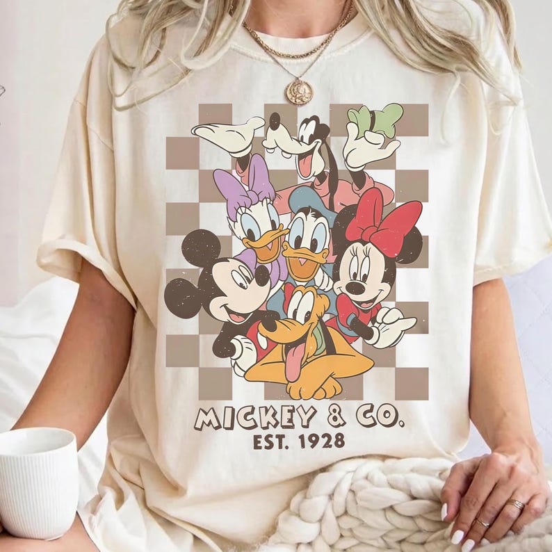 May include: Cream-colored t-shirt featuring a graphic of Mickey Mouse, Minnie Mouse, Donald Duck, Daisy Duck, Goofy, and Pluto. The design is set against a brown and white checkered background, with "Mickey & Co. Est. 1928" printed below.