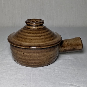 May include: A brown ceramic pot with a matching lid and a single side handle. The pot features a circular shape with horizontal ribbed textures along the body and the lid. The glaze has a speckled finish with darker brown tones throughout.