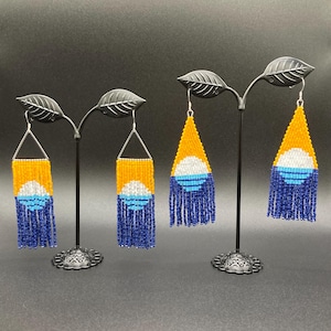May include: Handmade beaded earrings in a geometric design. The earrings feature a yellow top, white and blue center, and blue fringe. The earrings are displayed on black metal stands with leaf accents, against a dark background.