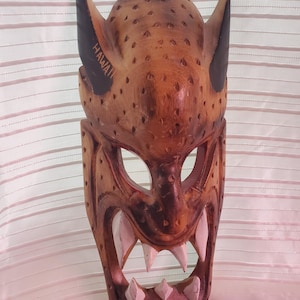 May include: A carved wooden mask with a fierce expression. The mask is brown with black accents on the horns and the word "HAWAII" carved on the side. The teeth are painted white.