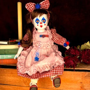 May include: A porcelain clown doll with a painted face, wearing a pink dress with a red and white checkered apron and sleeves. The doll has a matching bow and red shoes. A candle and books are in the background.