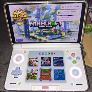 May include: A white handheld gaming console with a dual-screen display. The top screen shows the Minecraft Pocket Edition title screen and a Mythical Trainers Hub logo. The bottom touchscreen displays a grid of game icons including Mario and Minecraft.