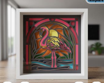 May include: A layered paper art piece featuring a pink flamingo in a stained-glass style design. The artwork is set against a sunset backdrop with a yellow sun and green foliage. The piece is framed in a white square frame. Text includes "+Bonus Frame Template", "SVG", "DXF", "YOUR 3D SVG", "PDF", and "PNG".