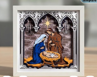 May include: A layered paper art piece depicting the Nativity scene, framed in white. Mary in blue and Joseph in brown gaze at the baby Jesus in a manger. The background features an arched structure and a star.