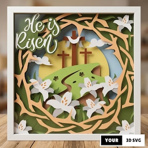 Puede incluir: Una obra de arte en papel en capas en un marco blanco, con un tema religioso de Pascua. El diseño incluye tres cruces, lirios blancos y el texto "He is Risen". El fondo tiene un sol amarillo y una colina verde.