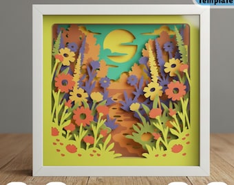 May include: A framed 3D paper art piece depicting a colorful landscape with flowers, trees, and a sun. The artwork features layers of paper cut into intricate shapes, creating a depth effect. The frame is white, and the overall design is vibrant and detailed.