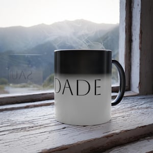 Könnte beinhalten: Eine farbwechselnde Tasse in Weiß und Schwarz mit dem Wort "DADE" aufgedruckt. Die Tasse steht auf einer verwitterten, weißen Holzoberfläche in der Nähe eines Fensters. Dampf steigt aus der Tasse auf, was auf ein heißes Getränk hindeutet.