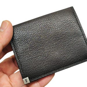 Peut inclure: Un portefeuille en cuir noir avec une surface textur&eacute;e. Le portefeuille est de forme carr&eacute;e et pr&eacute;sente des d&eacute;tails m&eacute;talliques argent&eacute;s aux coins. Le portefeuille est tenu dans une main.