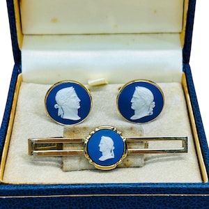 Op de afbeelding: Een vintage sieradenset met een stropdasklem en twee manchetknopen, gepresenteerd in een blauwe doos. Elk stuk heeft een goudkleurig frame met een blauw midden, met een wit camee-profiel. De set is waarschijnlijk uit het midden van de 20e eeuw.