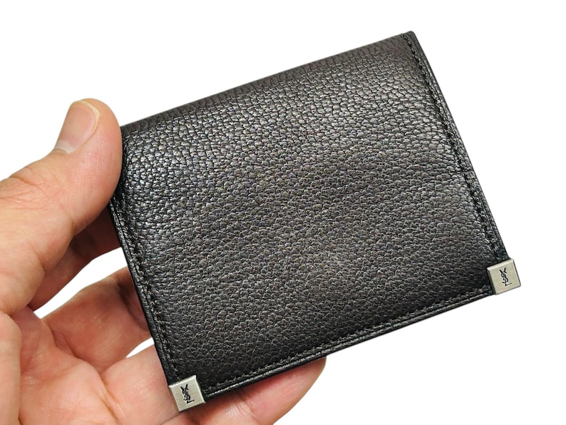Peut inclure: Un portefeuille en cuir noir avec une surface textur&eacute;e. Le portefeuille est de forme carr&eacute;e et pr&eacute;sente des d&eacute;tails m&eacute;talliques argent&eacute;s aux coins. Le portefeuille est tenu dans une main.