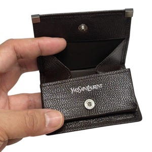 Peut inclure: Un porte-monnaie en cuir marron fonc&eacute; avec une fermeture &agrave; bouton-pression et des d&eacute;tails argent&eacute;s. L'int&eacute;rieur est marron clair. Le nom de la marque "Yves Saint Laurent" est visible en lettres blanches.