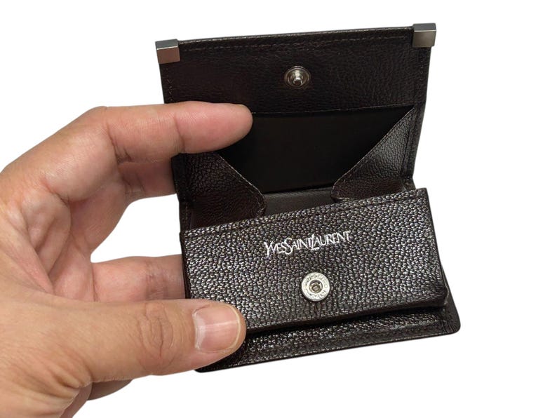 Peut inclure: Un porte-monnaie en cuir marron fonc&eacute; avec une fermeture &agrave; bouton-pression et des d&eacute;tails argent&eacute;s. L'int&eacute;rieur est marron clair. Le nom de la marque "Yves Saint Laurent" est visible en lettres blanches.
