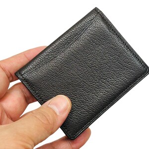 Peut inclure: Un porte-cartes en cuir noir avec une surface textur&eacute;e. Le porte-cartes rectangulaire a des coins arrondis et une bordure cousue. Il est tenu dans une main sur fond blanc.