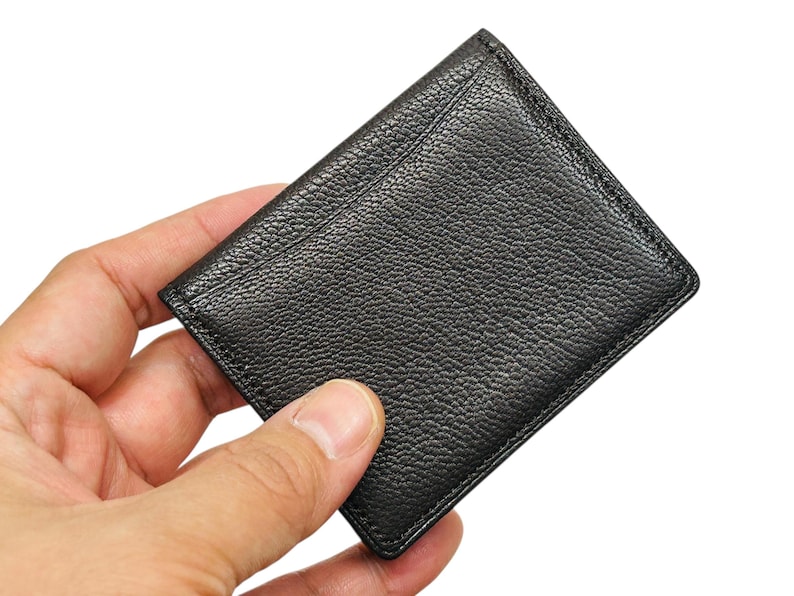Peut inclure: Un porte-cartes en cuir noir avec une surface textur&eacute;e. Le porte-cartes rectangulaire a des coins arrondis et une bordure cousue. Il est tenu dans une main sur fond blanc.