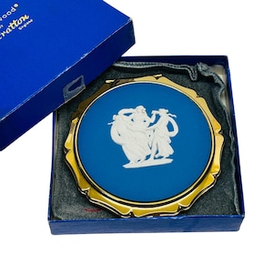 May include: A blue compact mirror with a gold-colored frame and a white relief design of figures. The mirror is in a blue box with the text "Wedgwood by Stratton England". The compact is a vintage collectible.