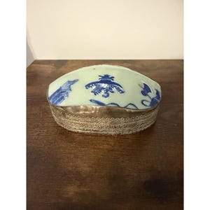 Puede incluir: Una caja ovalada con tapa de cerámica verde pálido decorada con motivos pintados en azul. La base del recipiente está fabricada en metal con un intrincado patrón de filigrana en relieve que rodea todo el perímetro del objeto.
