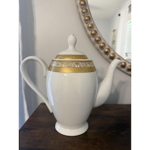 May include: A white porcelain teapot featuring a gold band around the upper body and lid. The teapot has a curved handle, a tapered spout, and a matching lid with a small knob handle. The gold band displays a detailed, intricate pattern.