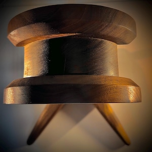 May include: A handcrafted wooden stool with a dark brown finish. The stool features a round seat, a cylindrical body, and three angled legs. The design is minimalist and modern, suitable for various interior styles.