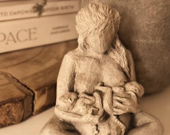 Op de afbeelding: Een neutrale sculptuur van een zittende figuur die twee baby's borstvoeding geeft. De sculptuur heeft een getextureerd, gebeeldhouwd uiterlijk en staat tegen een vage achtergrond met boeken en een groot, afgerond object.