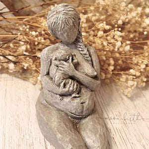 Puede incluir: Una escultura gris de una madre sosteniendo a un bebé. La madre está arrodillada, con la cabeza inclinada, y su cabello está trenzado. La escultura tiene una apariencia texturizada y está colocada sobre un fondo borroso de flores secas.